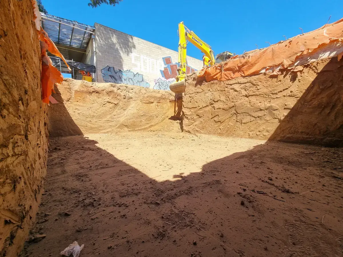 Deep bulk excavation with vertical cut walls, an excavator working from the top edge beside a commercial building