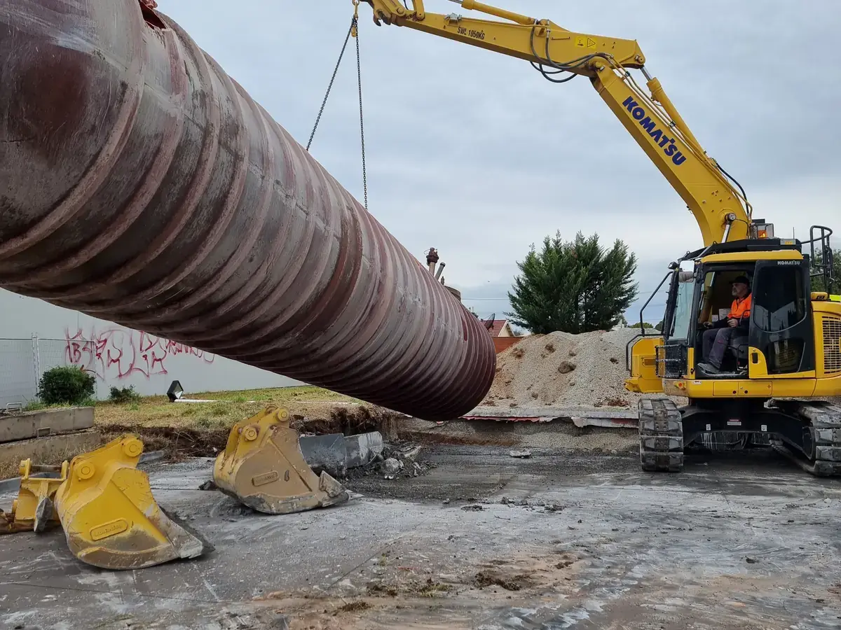 Excavator lifting a large ribbed steel underground storage tank out of the ground on a cleared commercial site